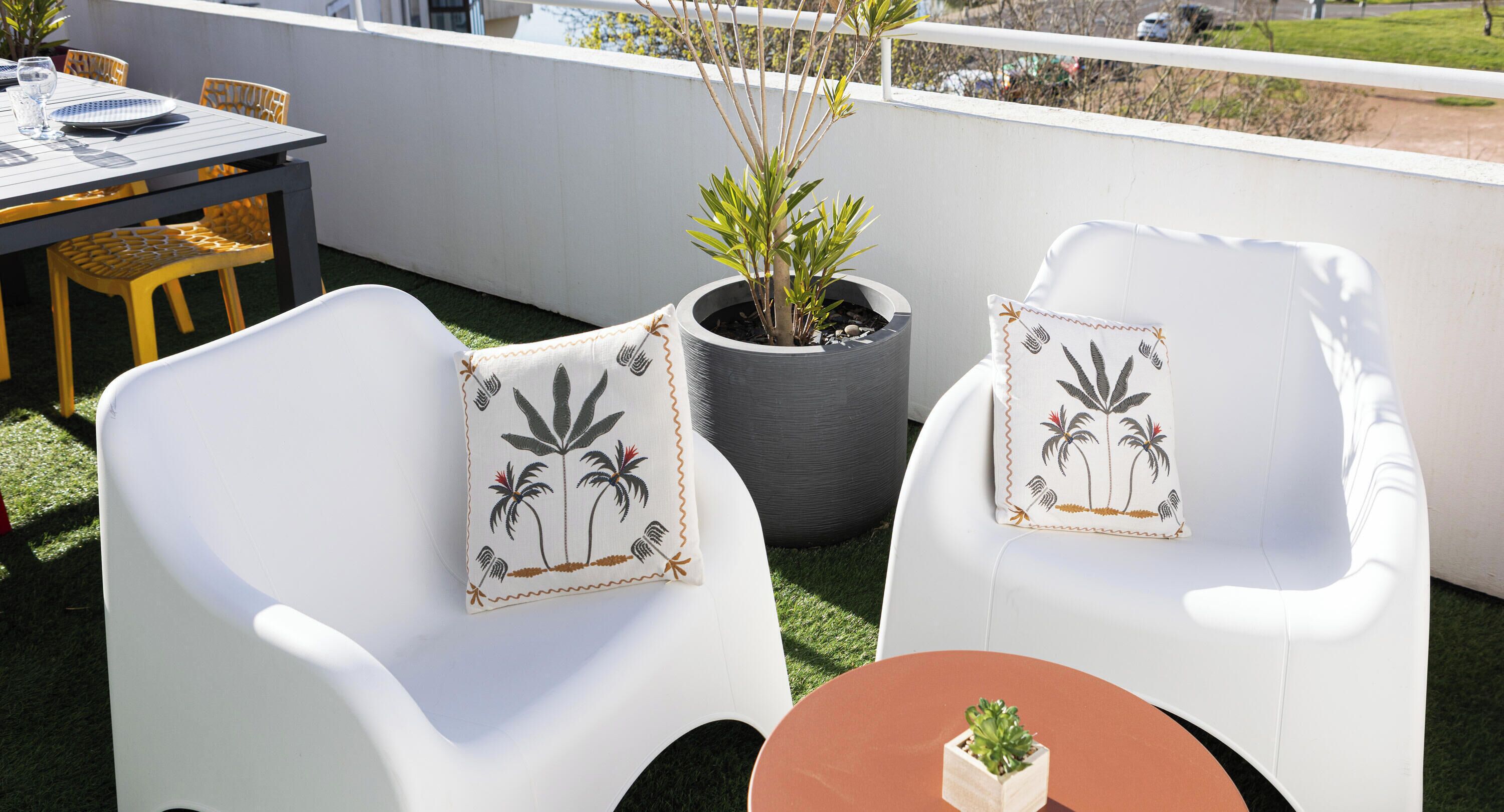 Two white chairs with matching cushions, a small table, and a potted plant on an outdoor patio.