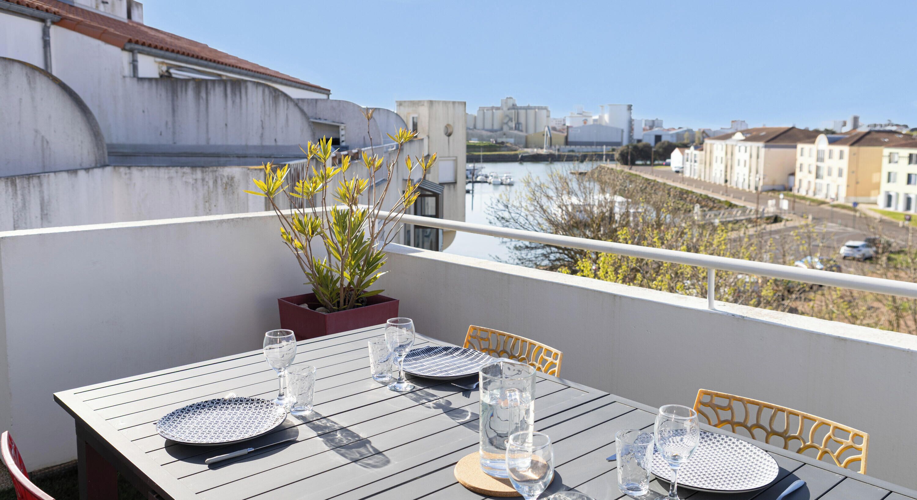 A balcony with a table set for dining, a potted plant, and a view of a river and buildings.