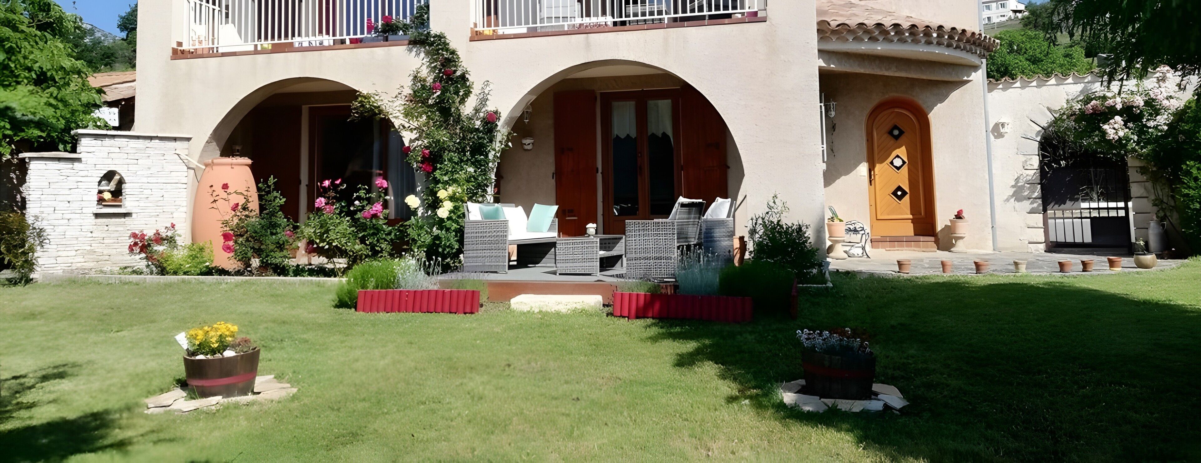 Un bâtiment doté de deux entrées en arc de cercle, d’un balcon avec des balustrades blanches et d’un jardin agrémenté de plantes en pot et de mobilier d’extérieur.