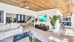 A spacious living room with a large flat-screen TV, a ceiling fan, and a modern kitchen area. - Spacious Tropical Pool Home Miami. 4/3. Sleeps 13. Large Garden/ Backyard. (Miami)