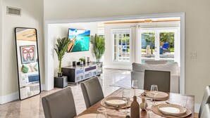 A dining area with a wooden table set for four, a TV mounted on the wall, and a view of the outdoors through a glass door. - Spacious Tropical Pool Home Miami. 4/3. Sleeps 13. Large Garden/ Backyard. (Miami)