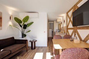 Ein modernes Wohnzimmer mit einer Couch, einem Esstisch und Stühlen. Eine große Pflanze, ein kleiner Beistelltisch und ein Flachbildfernseher, der an einer Holzwand angebracht ist, sind ebenfalls vorhanden.