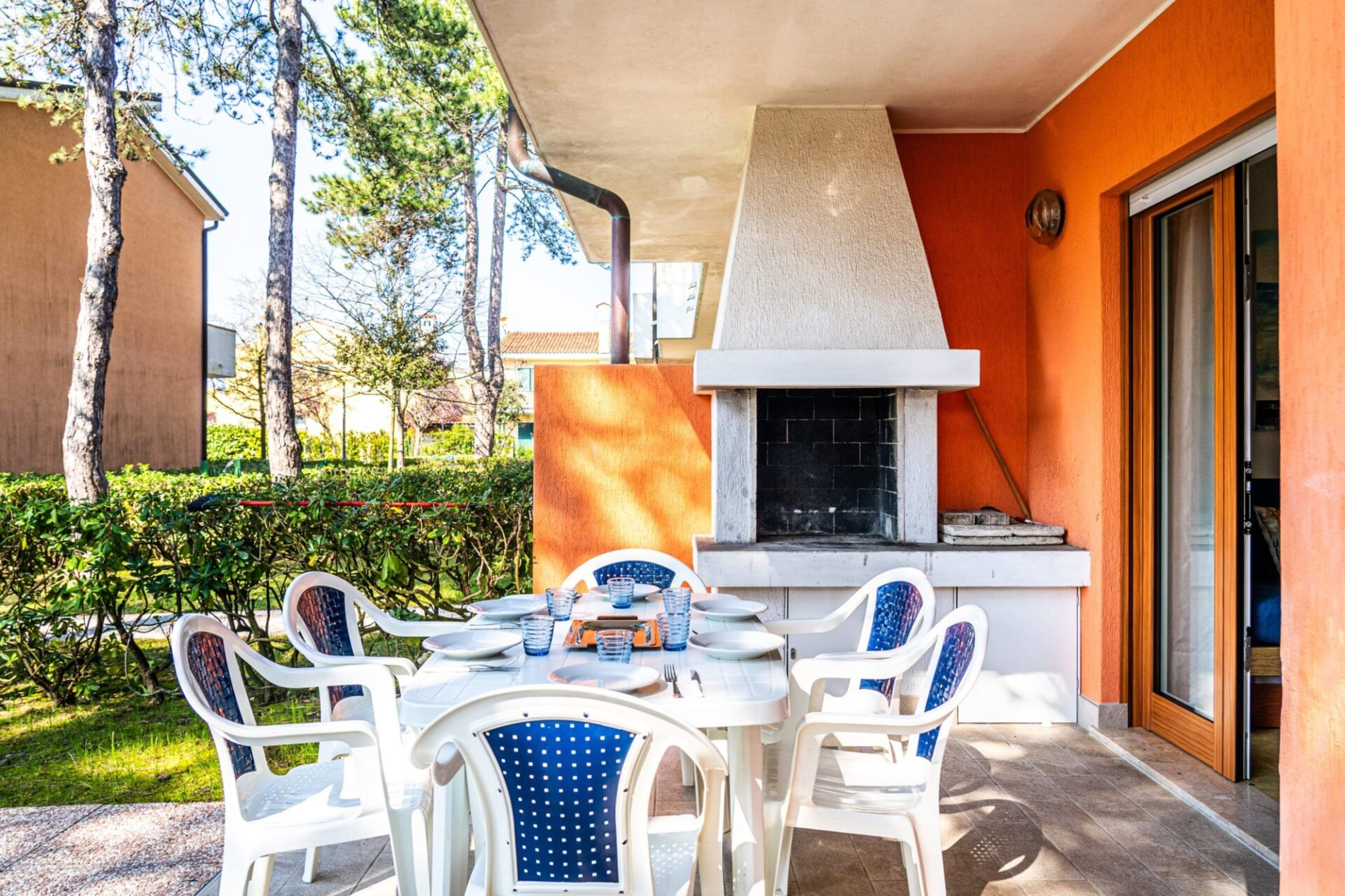 An outdoor patio with white plastic chairs and a table set for dining, a barbecue grill, and a view of the surrounding greenery.