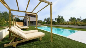 Un espace aménagé au bord de la piscine, avec une pergola en bois, deux chaises longues et une maison moderne en arrière-plan.