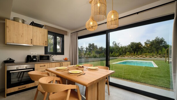 Une cuisine moderne avec des meubles en bois, une table à manger avec des chaises, un four encastré et une vue sur une piscine à travers de grandes fenêtres.