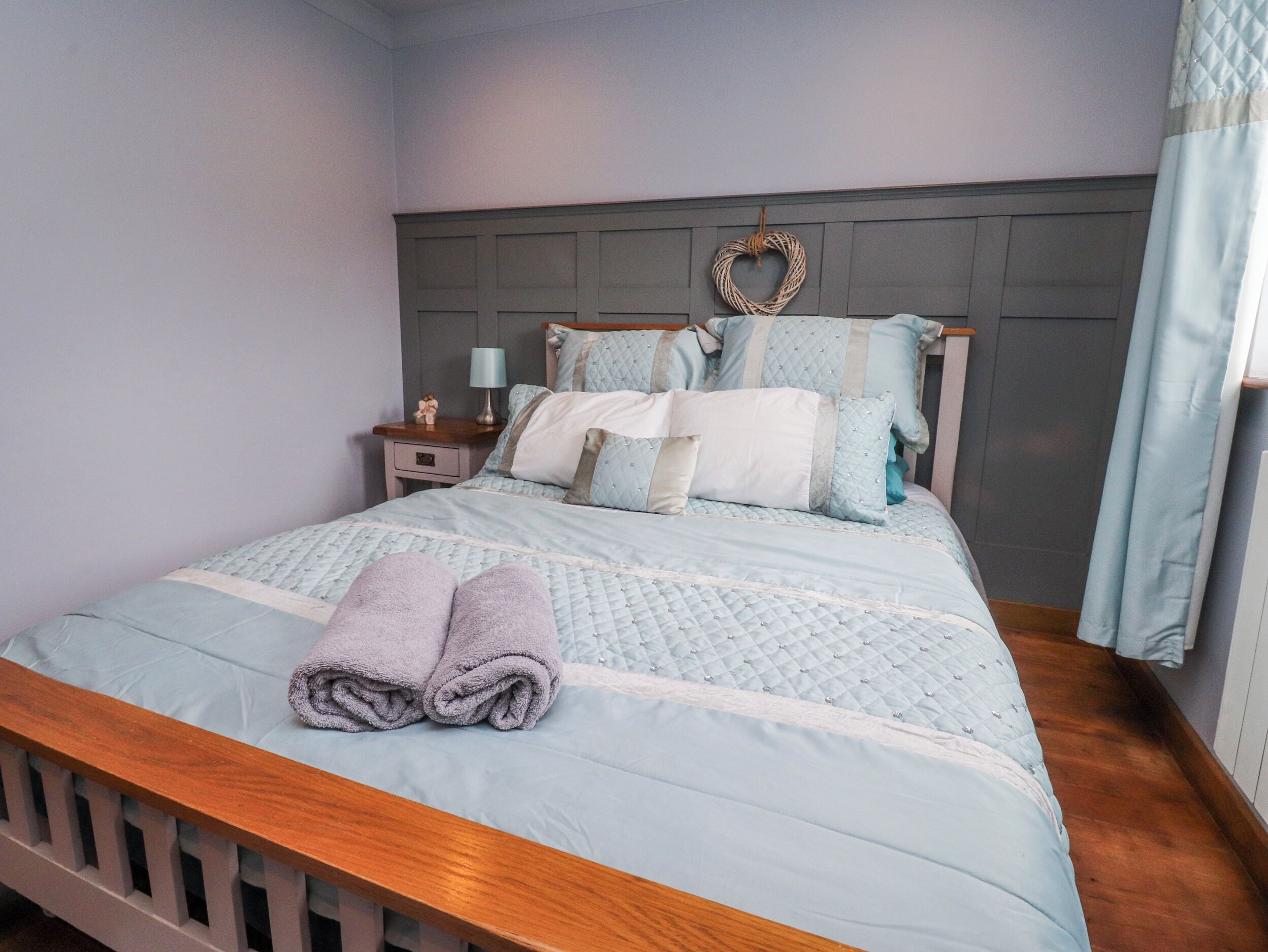 Un lit bien fait, avec une tête de lit, une table de chevet avec une lampe et une décoration en forme de cœur accrochée au mur.