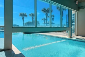 A swimming pool with clear blue water, large windows, and palm trees in the background. - Tradewinds #1108 - Beachfront 1BR + Bunks, Gulf View, Indoor/Outdoor Pool (Orange Beach)
