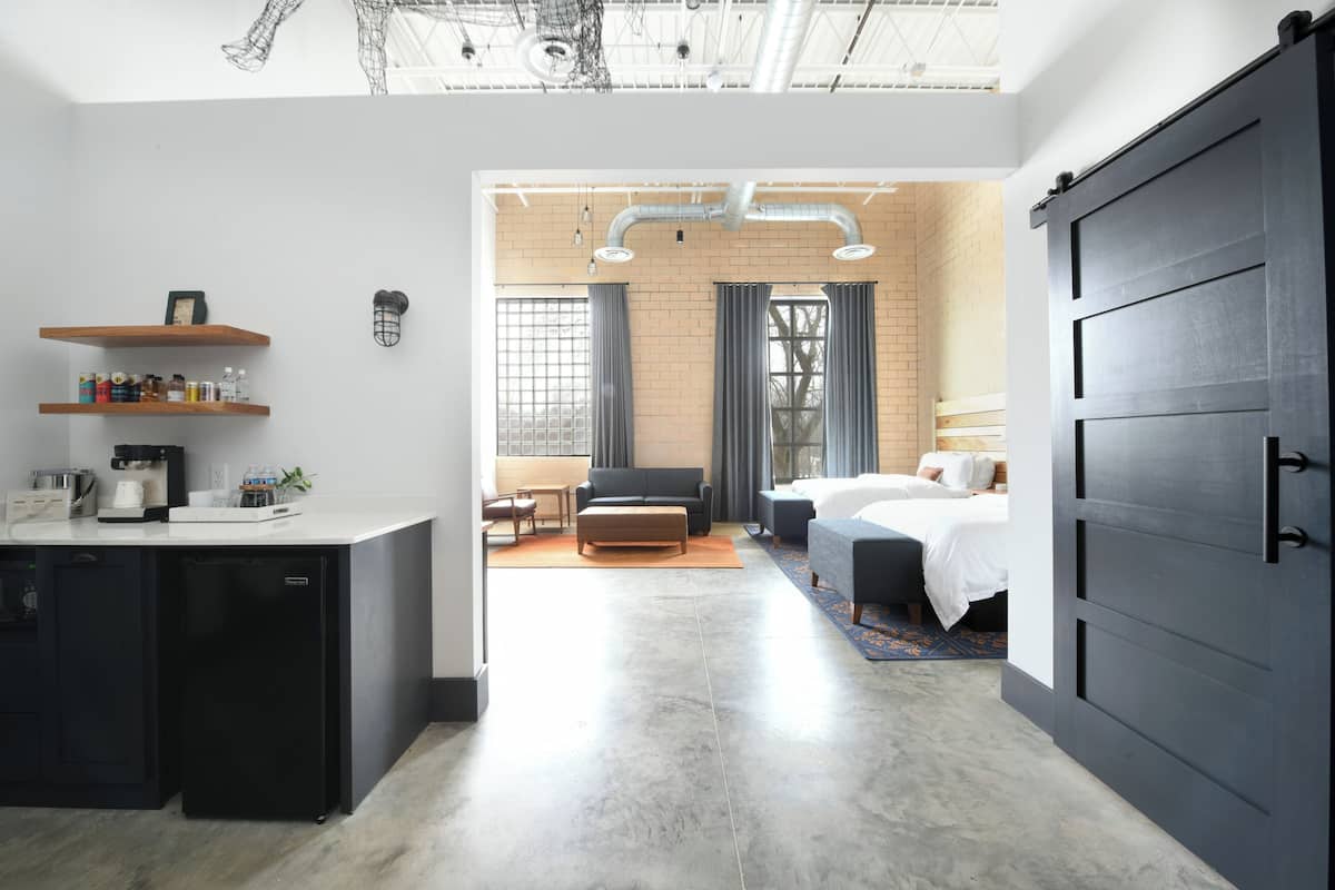 A modern hotel room with a kitchen area, a dining table, and a large bed.