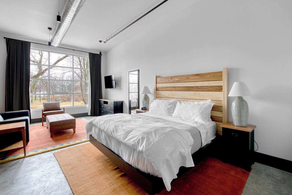 A modern hotel room with a large bed, a wooden headboard, a flat-screen TV, a bedside table with a lamp, and a view of trees through the window.