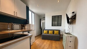 Een compacte keuken met moderne apparaten, een grijze bank met gele kussens en een flatscreen televisie aan de muur.
