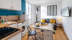 Een knus woongedeelte met kitchenette, een eethoek, een bank met kussens, een televisie en een wasmachine.
