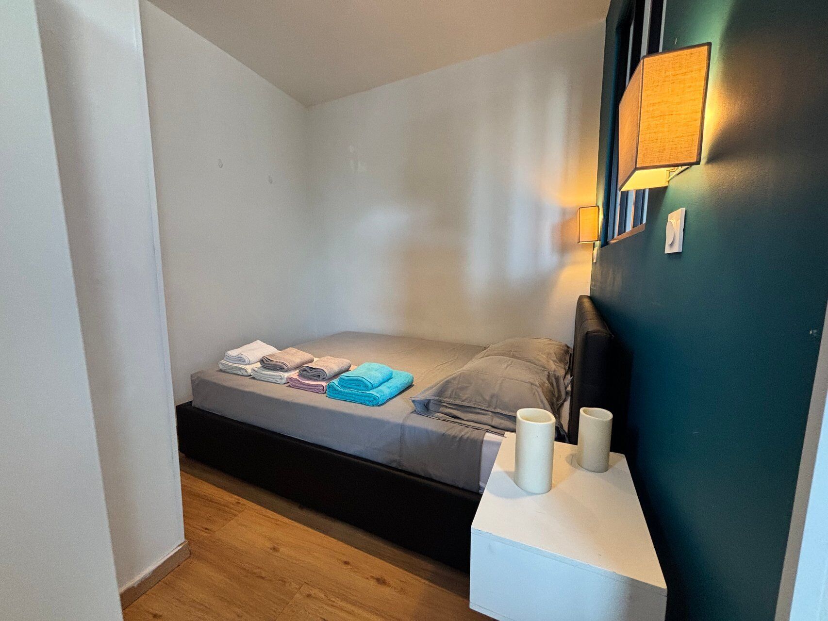 A small, narrow room with a bed, a nightstand, and a wall-mounted lamp.