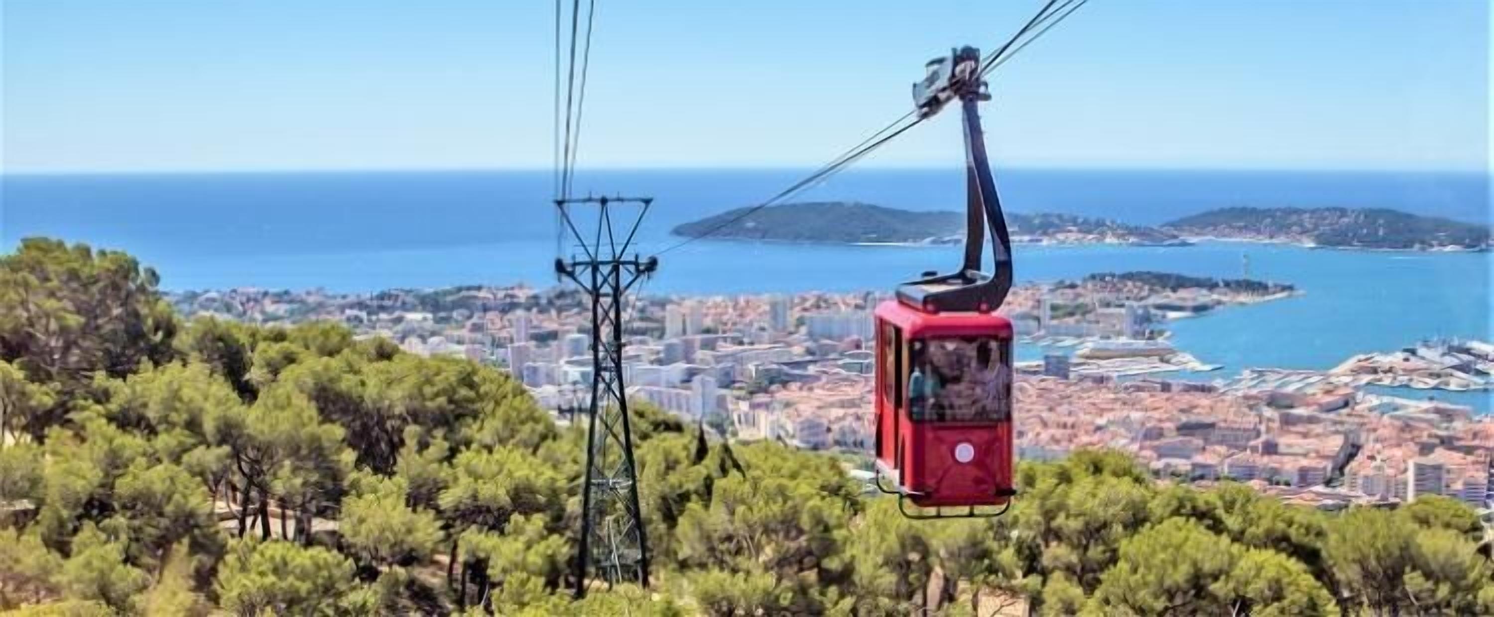 Un teleférico con cabina roja, un paisaje urbano y la costa al fondo.