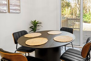 Une table ronde, surmontée de quatre sets de table en fibres tressées, entourée de chaises noires agrémentées d’éléments en bois, le tout placé devant un mur blanc orné de deux œuvres d’art encadrées.