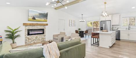 A modern living room with a fireplace, green sofa, dining area, kitchen, and a large window.