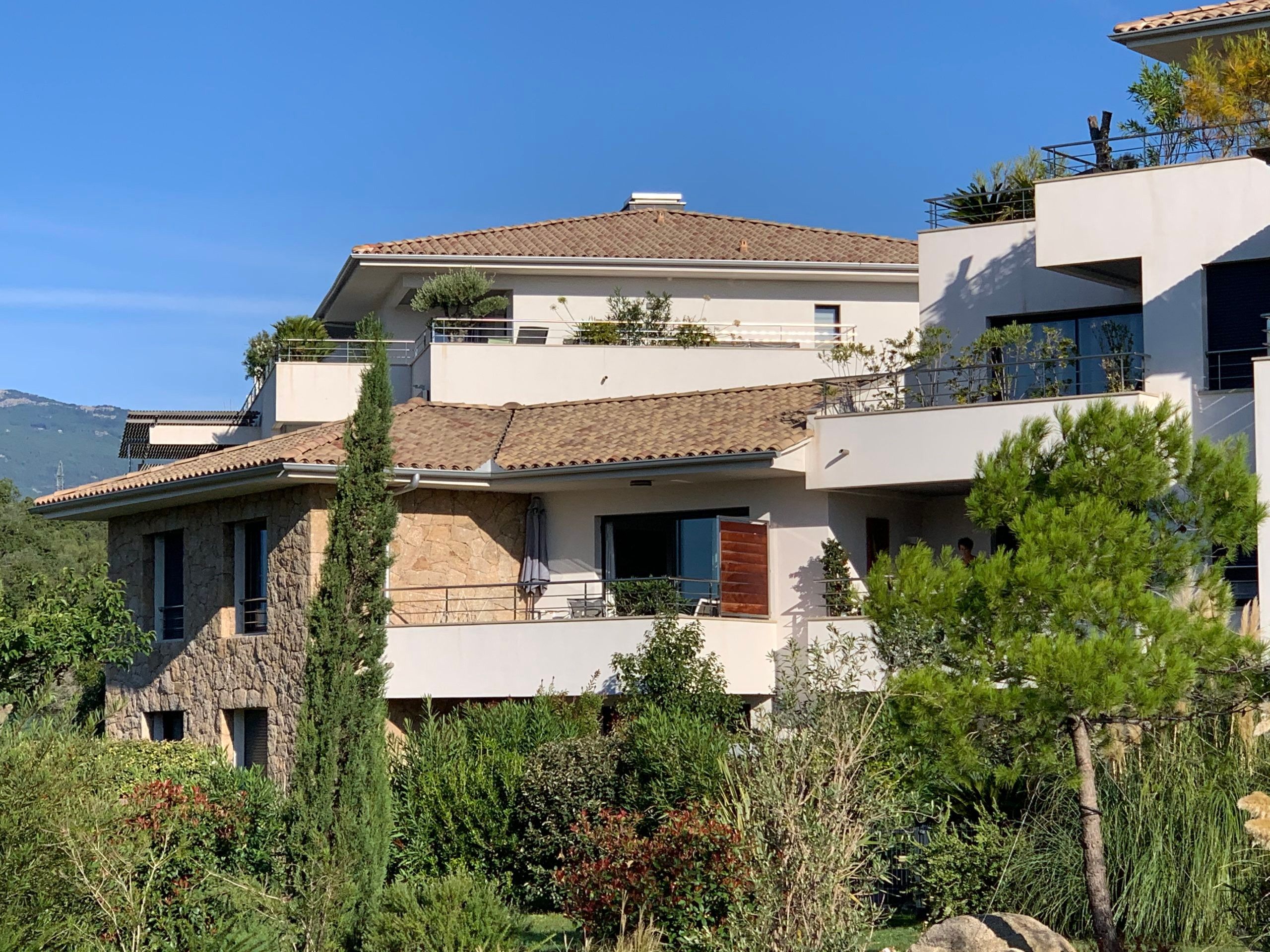 A multi-story residential building with a stone facade, balconies, and a view of the surrounding greenery and mountains.