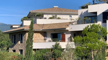 A multi-story residential building with a stone facade, balconies, and a view of the surrounding greenery and mountains.