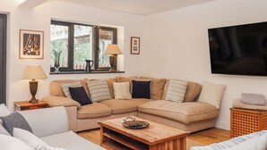A living room with a sectional sofa, a wooden coffee table, a TV mounted on the wall, and a window with a view of greenery. - Lower Moorwood (Newton Abbot)