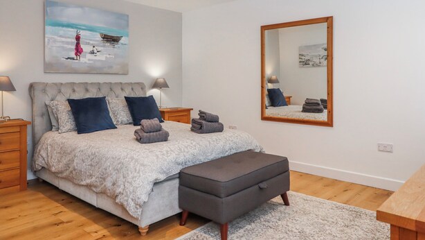 A bedroom with a bed, bedside tables, a bench, a large mirror, and artwork on the wall. - Lower Moorwood (Newton Abbot)