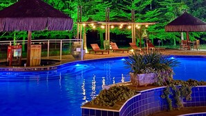 A swimming pool with a tiled edge, surrounded by lounge chairs and thatched umbrellas, set against a backdrop of dense foliage.