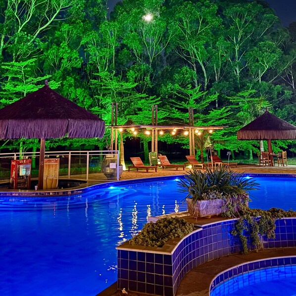 A swimming pool with a tiled edge, surrounded by lounge chairs and thatched umbrellas, set against a backdrop of dense foliage. - Sitio Castelinho Hotel Fazenda (Cesário Lange)