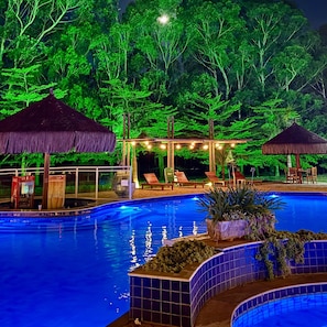A swimming pool with a tiled edge, surrounded by lounge chairs and thatched umbrellas, set against a backdrop of dense foliage. - Sitio Castelinho Hotel Fazenda (Cesário Lange)