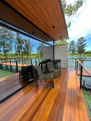 A wooden deck with outdoor furniture, a brick wall, and a view of a lake and trees. - Sitio Castelinho Hotel Fazenda (Cesário Lange)