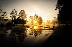 A bridge over a calm body of water with trees on either side and a sunrise in the background. - Sitio Castelinho Hotel Fazenda (Cesário Lange)
