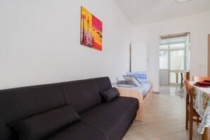 Ein modernes Zimmer mit einem schwarzen Sofa, einem Bett mit blauer und weißer Bettdecke, einem Holztisch mit floralem Tischläufer und einem farbenfrohen, abstrakten Gemälde an der Wand.