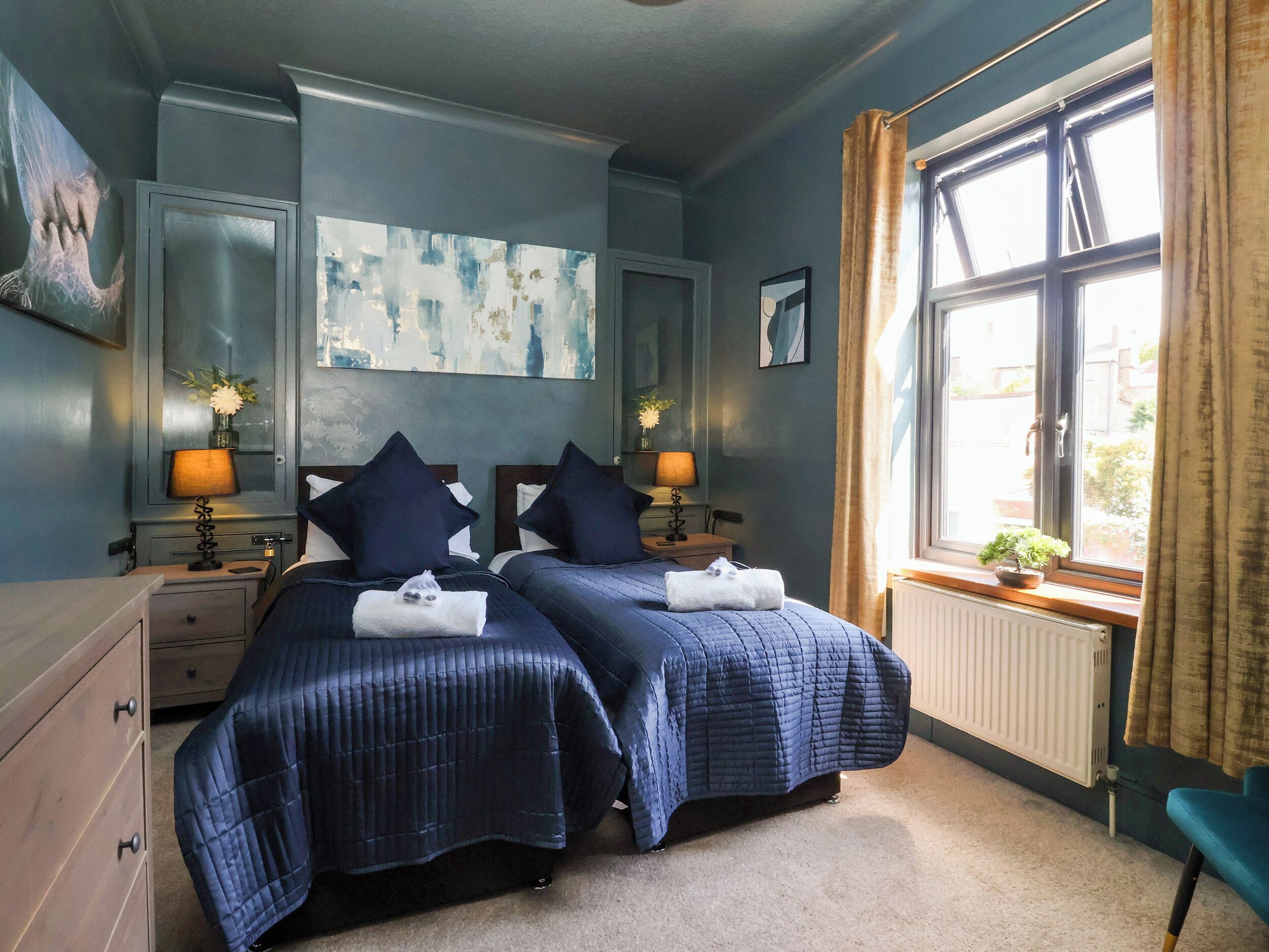 A hotel room with two beds, a large window with curtains, a radiator, and artwork on the walls.