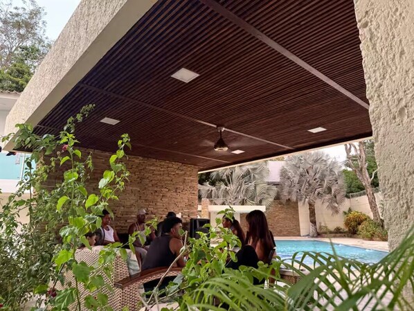 Un área de estar al aire libre con piscina, cubierta por un techo de madera, rodeada de vegetación.