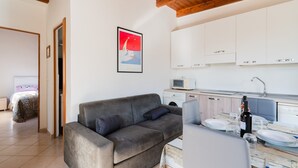 Una sala moderna con un sofá gris, una mesa de comedor con platos y una botella, una cocina con armarios blancos y un cuadro en la pared.