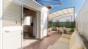 Un patio descubierto con un sofá blanco, una mesa de comedor con sillas y una cocina visible a través de una puerta abierta.