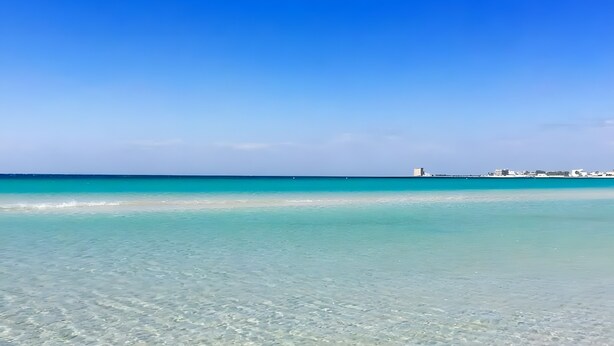 Una playa con agua turquesa cristalina, una orilla arenosa y un edificio a lo lejos.