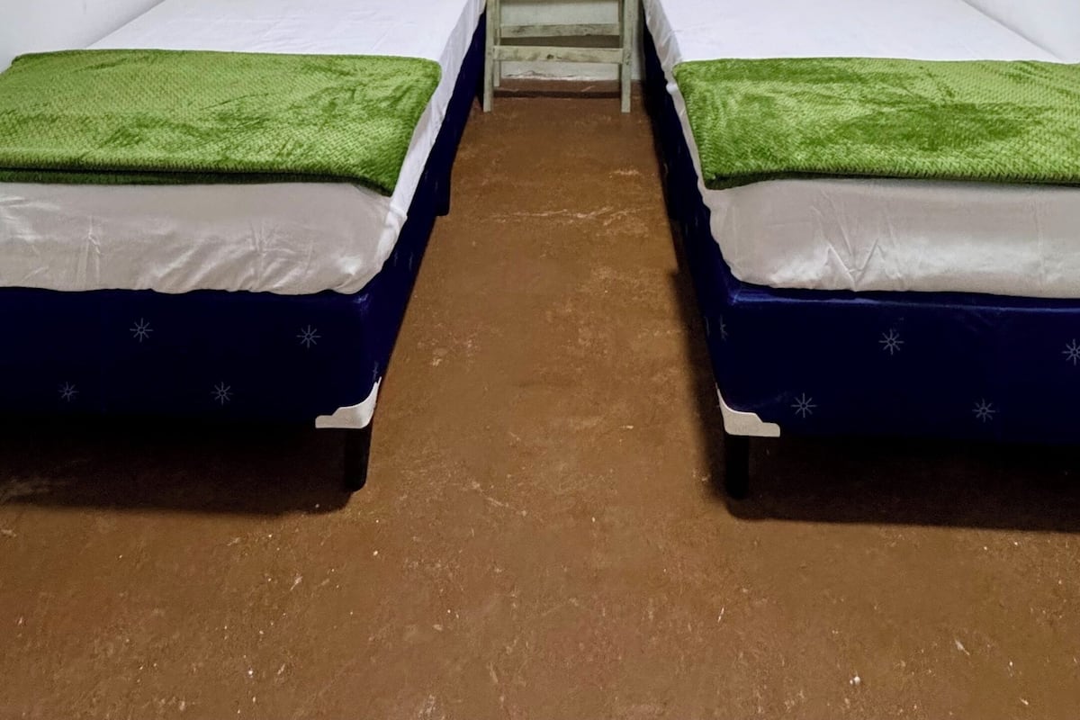 Una habitación pequeña con dos camas, una mesita de noche, una lámpara colgante y dos cuadros en la pared.