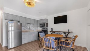 A compact kitchen with grey cabinets, a stainless steel refrigerator, a microwave, and a built-in oven. There is a round table with wicker chairs and a patterned rug. - Moonraker 15 (Gulf Shores)