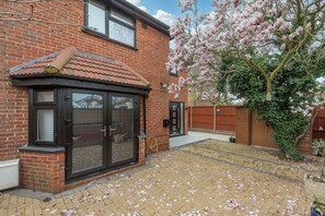 A brick house with a conservatory, a garden with a tree bearing pink flowers, and a paved driveway. - Luxurious 3BDR 3BATH Hounslow by the Airport (Greater London)
