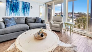 Ett vardagsrum med en grå soffa, ett runt soffbord och ett stort fönster med utsikt över hus.