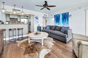 A modern living room with a grey sofa, a round coffee table, a ceiling fan, and a kitchen area with light green cabinets and a tiled backsplash. - Six Palms 3A (Santa Rosa Beach)
