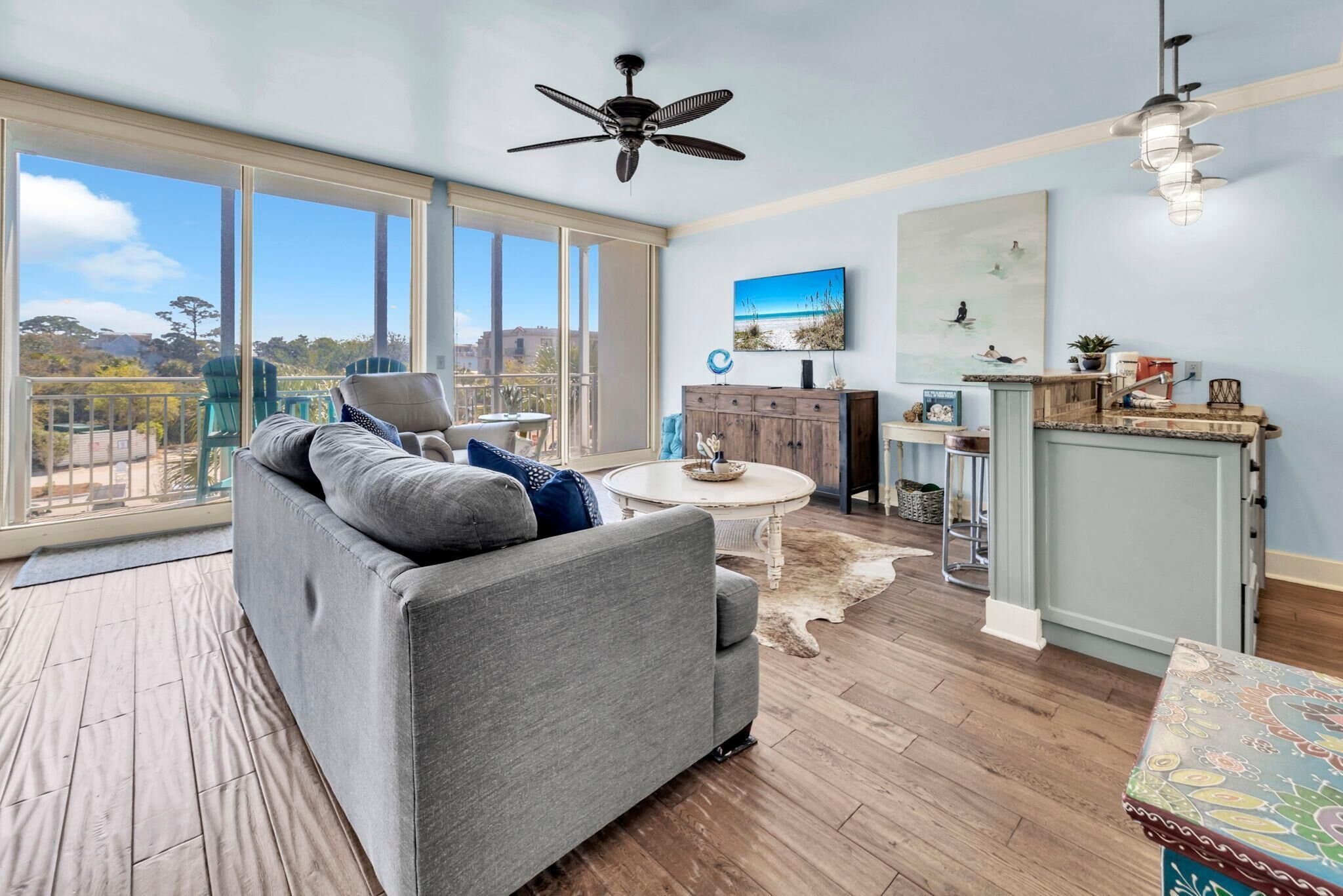 A spacious living room with a grey sofa, a ceiling fan, a dining table, and a view through large windows.