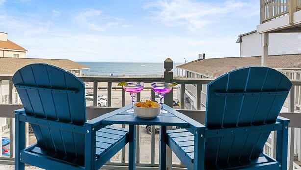 A balcony with two blue Adirondack chairs, a table with a bowl of chips, and two cocktails. - Twin View: 1 BR, Living Room, Ocean & Marsh Views, Beach Chairs, 1 parking spot (Hampton)