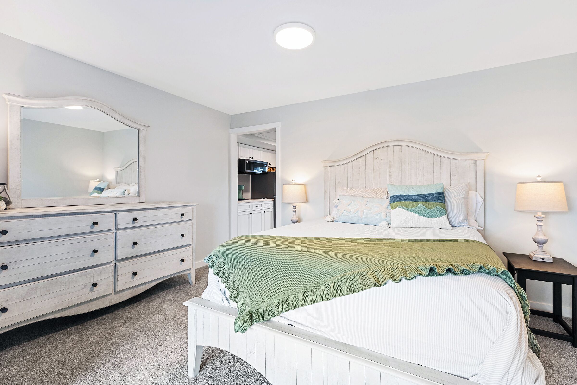 A bedroom with a white dresser, a bed with a green blanket, two bedside lamps, and a large mirror. - Twin View: 1 BR, Living Room, Ocean & Marsh Views, Beach Chairs, 1 parking spot (Hampton)
