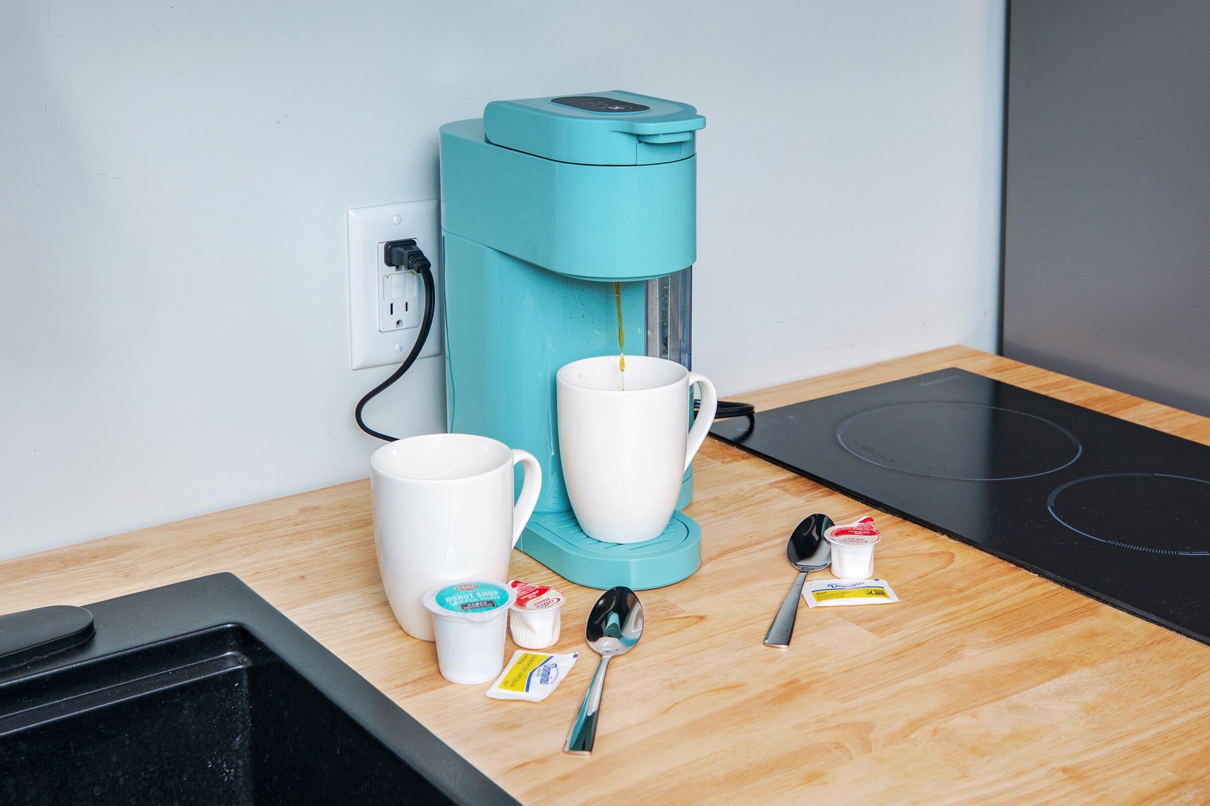 A teal coffee maker on a wooden countertop with two white mugs, a spoon, and a small container of cream.