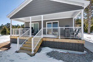 A covered porch with a wooden staircase, a white railing, and a view of a snowy landscape. - Steps to Lake Michigan | Hot Tub + Fire Pit | Near Cave Point (Sturgeon Bay)
