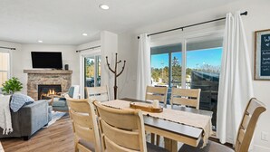 Un salon moderne avec une cheminée, un coin repas avec une table et des chaises, un téléviseur à écran plat fixé au mur et une grande fenêtre avec des rideaux.