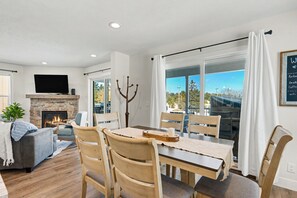 A modern living room with a fireplace, a dining area with a table and chairs, a flat-screen TV mounted on the wall, and a large window with curtains. - Steps to Lake Michigan | Hot Tub + Fire Pit | Near Cave Point (Sturgeon Bay)
