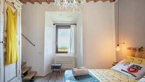 Ein Schlafzimmer mit einem Bett, einem Fenster mit Vorhängen, einem Heizkörper, einem kleinen Hocker und einer Deckenleuchte.