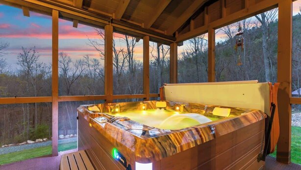 Een hottub met een houten buitenkant, een bankje en uitzicht op bomen door grote ramen.