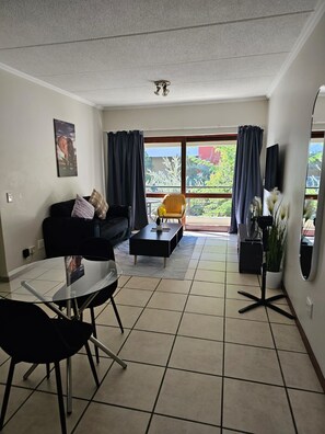 Ein Wohnzimmer mit Sofa, Couchtisch und Esstisch mit Stühlen. Es gibt einen Balkon mit Vorhängen und Blick auf Grünflächen.