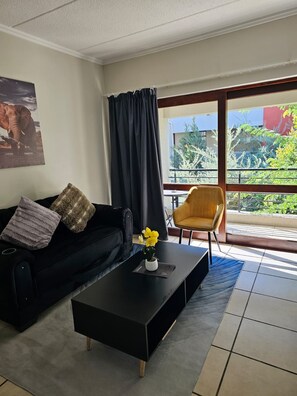 Ein Wohnzimmer mit einer schwarzen Couch, einem gelben Sessel, einem schwarzen Couchtisch mit einem Blumentopf und einem großen Fenster mit Blick auf Grünflächen und Gebäude.
